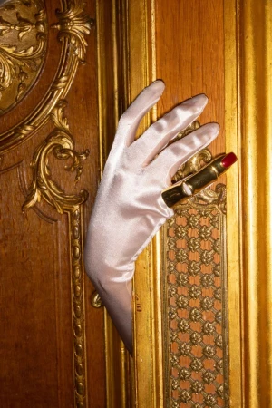 Elegant hand in satin glove holds gold lipstick against a luxurious gold-trimmed wooden panel, evoking classic beauty.