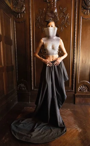 Model wearing a structured grid-patterned high collar top and long dark skirt inside a wood-paneled room