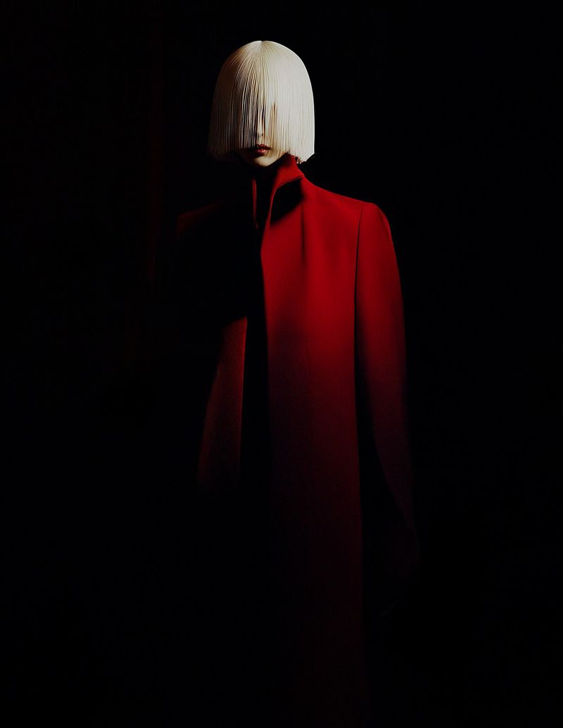 Fashionable woman with white fringe wig and red coat poses in dark studio for dramatic, high-fashion aesthetic in editorial style.