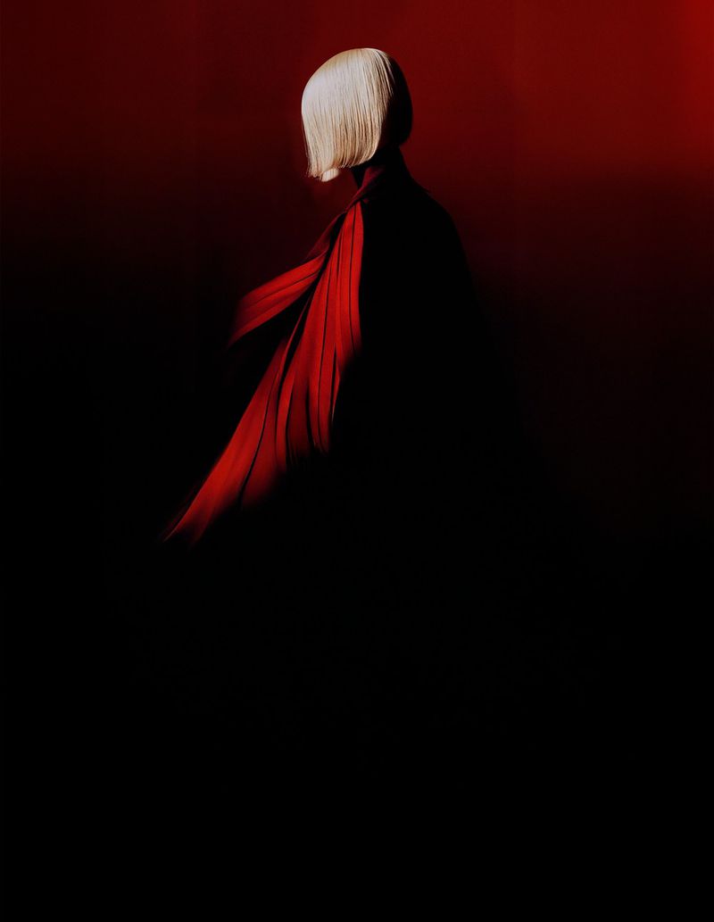 Side view of a person with a white bob haircut wearing a dark garment with a red fabric accent against a dark red gradient background.