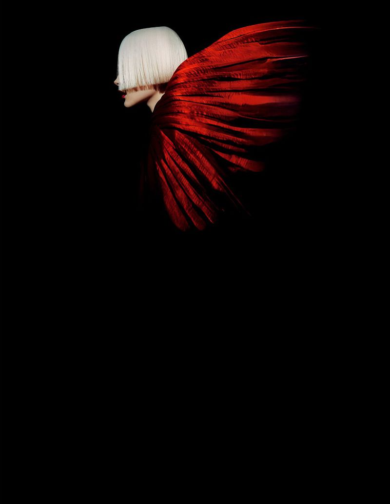 Profile of a person with a short white bob and red feathered wing on a solid black background in a studio lit portrait.