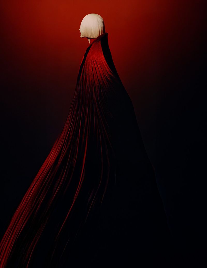 Person with white hair and long red flowing cape in a dark, dramatic portrait, showcasing fashion and visual artistry against a gradient background.