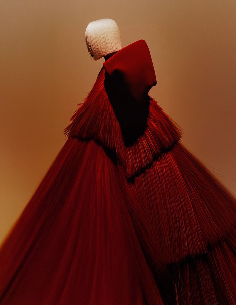 Fashion shot featuring a model in a textured, layered red gown and white bob hairstyle, set against a warm, golden-brown gradient background.