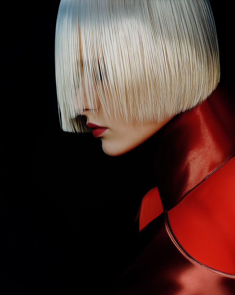 Close-up studio shot of a model with a blonde bob haircut with bangs, wearing red lipstick and a matching red metallic top, black background.