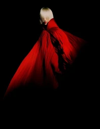 Model with platinum blonde blunt bangs wearing a voluminous red pleated cape against a black background.