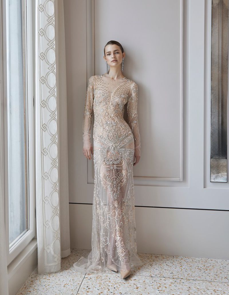 A woman wearing a formal gown poses in a bright interior space with neutral tones, featuring detailed architectural molding and a window.