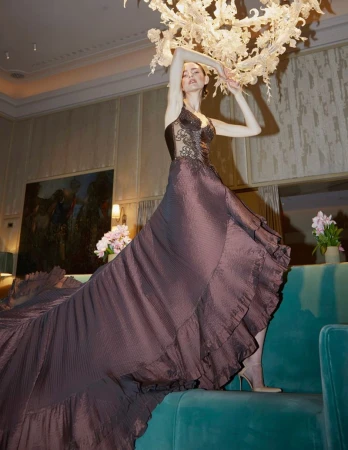 A slender adult female model poses in a flowing, dark-brown halter gown with lace details under an ornate chandelier in an indoor setting.