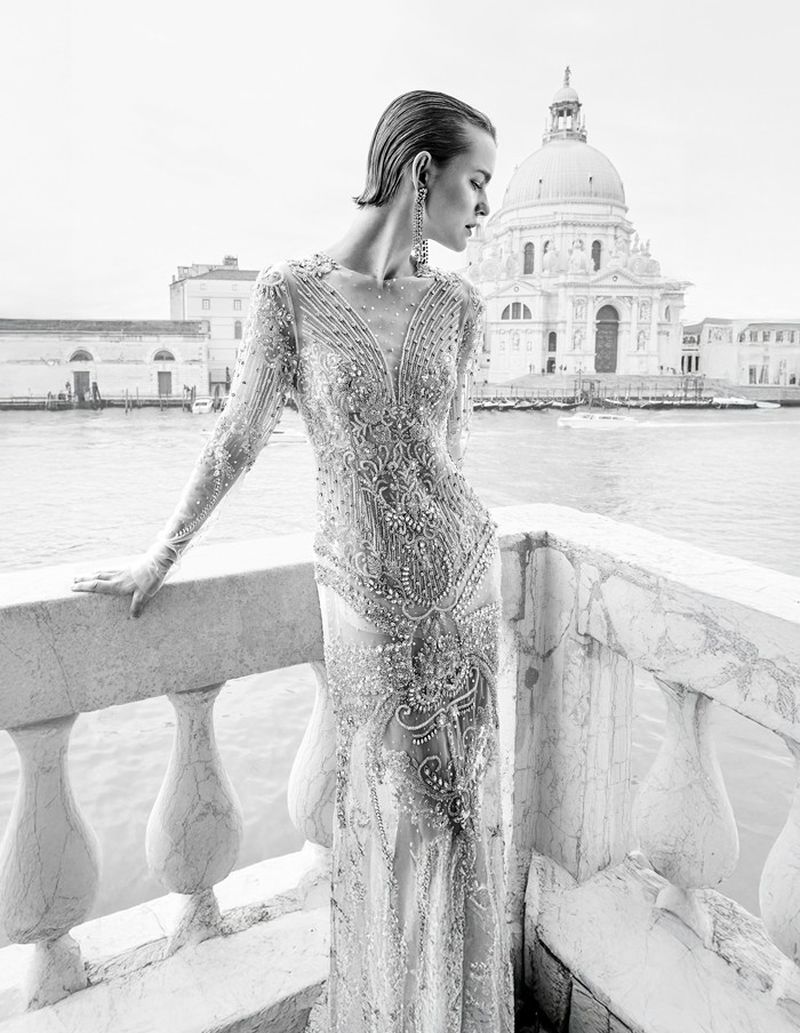 Monochrome image of a woman in an embellished gown on a balcony with a view of a domed building and water.
