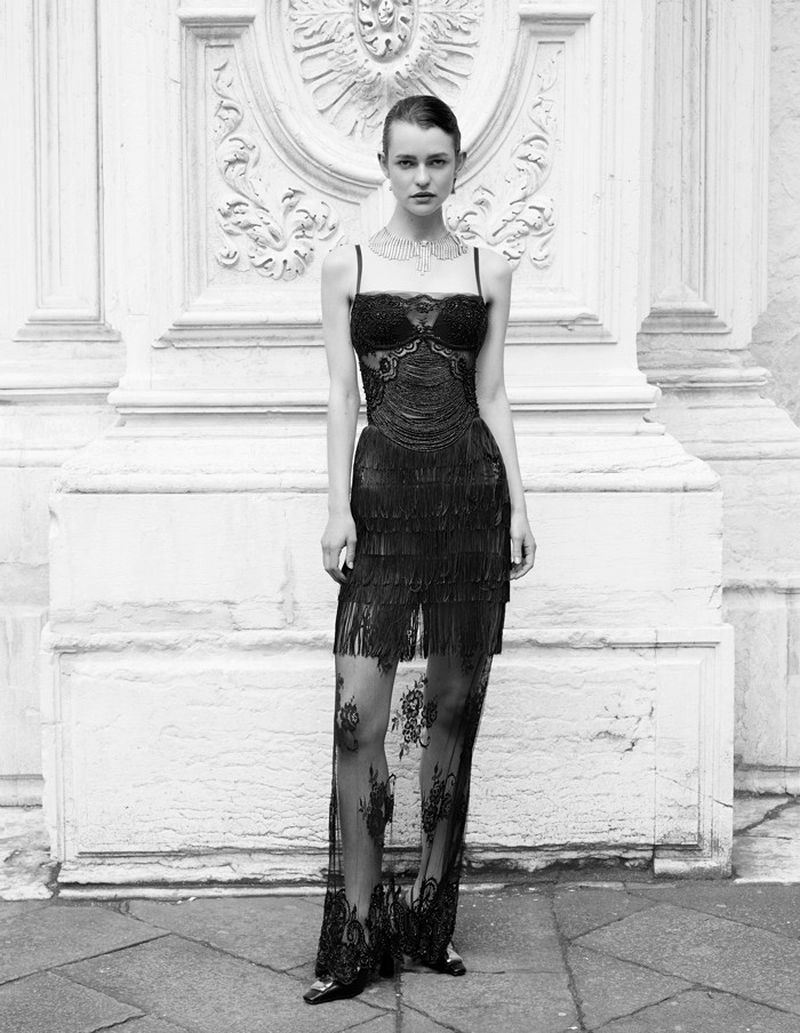 A fashion model poses in a black dress with fringe and lace details against a white ornate wall in an eye-level medium shot.
