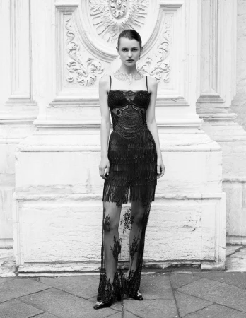 A fashion model poses in a black dress with fringe and lace details against a white ornate wall in an eye-level medium shot.