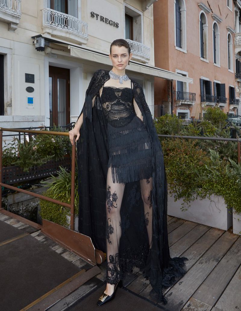 Fashionable adult female model poses in black lace dress with cape and jewelry on a bridge in Venice, Italy with hotel background.