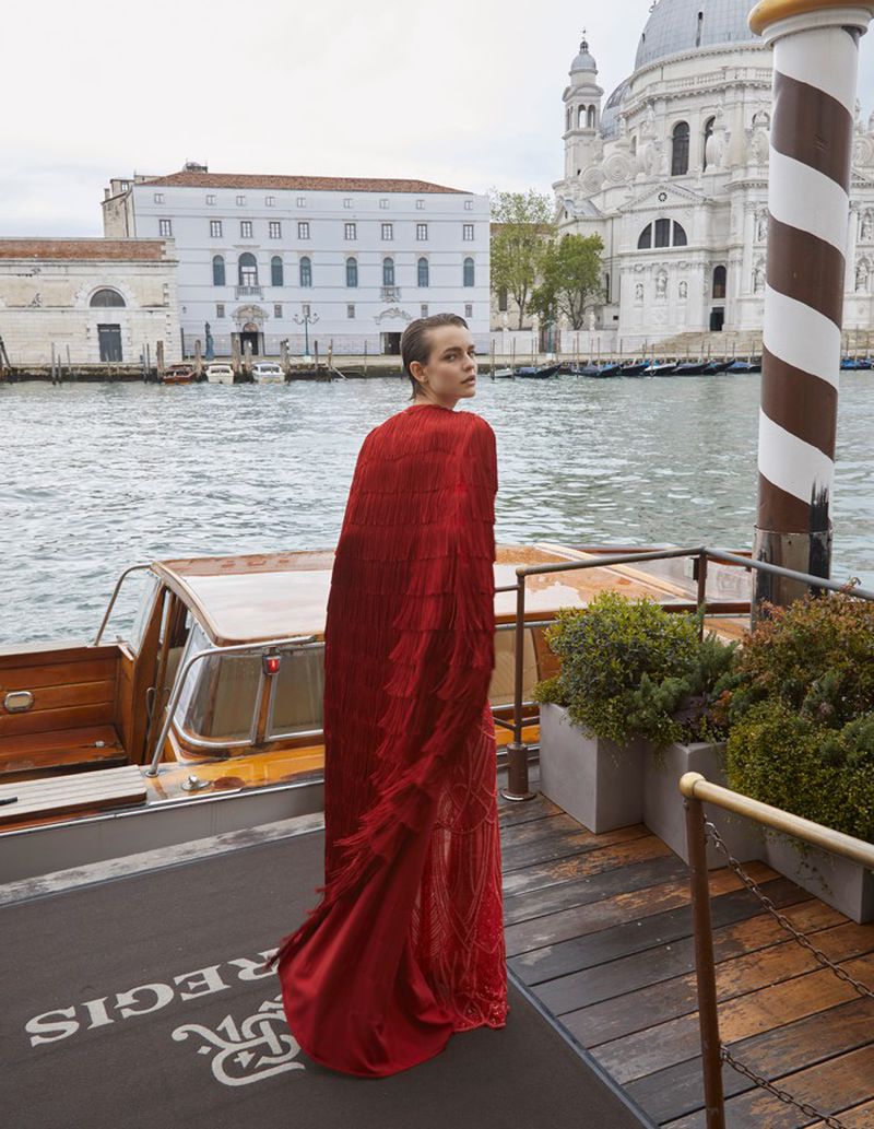 Woman in a red gown and cape standing on a dock in Venice, Italy, with a water taxi, building, and canal visible in the background.