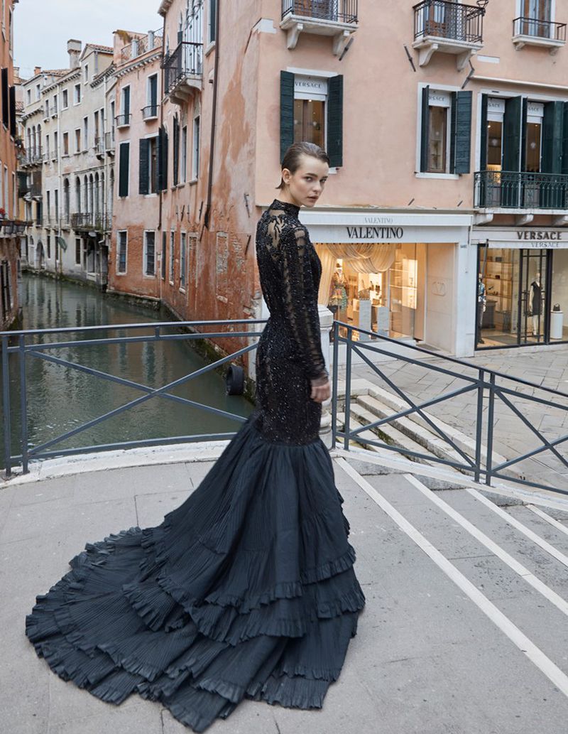 Fashionable adult woman in a black gown poses on a bridge in Venice near a canal. Storefronts and buildings in the background.