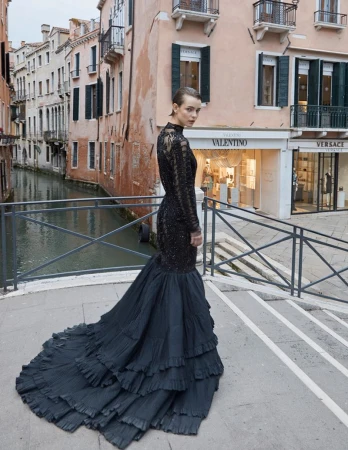 Fashionable adult woman in a black gown poses on a bridge in Venice near a canal. Storefronts and buildings in the background.