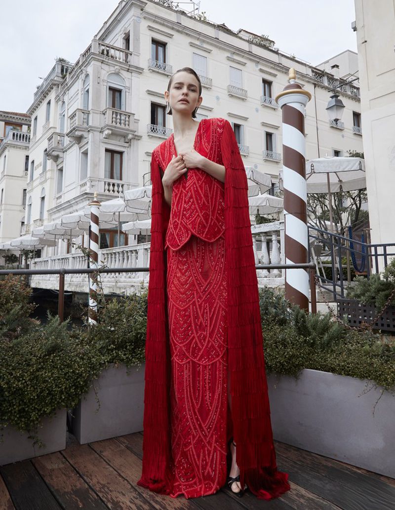 Elegant woman poses in a red beaded cape dress on a balcony with Venetian architecture in the background, showcasing fashion and style.