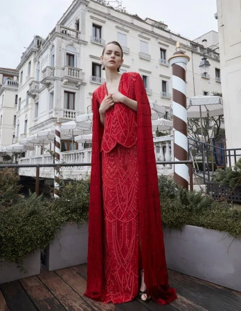 Elegant woman poses in a red beaded cape dress on a balcony with Venetian architecture in the background, showcasing fashion and style.