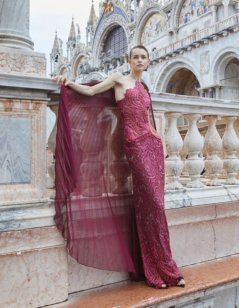 A model in a magenta gown poses on a stone balcony with ornate architecture behind her during a fashion shoot.