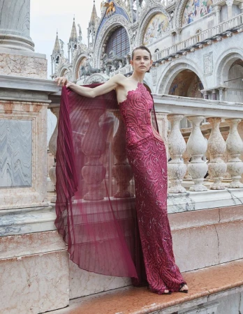 A model in a magenta gown poses on a stone balcony with ornate architecture behind her during a fashion shoot.