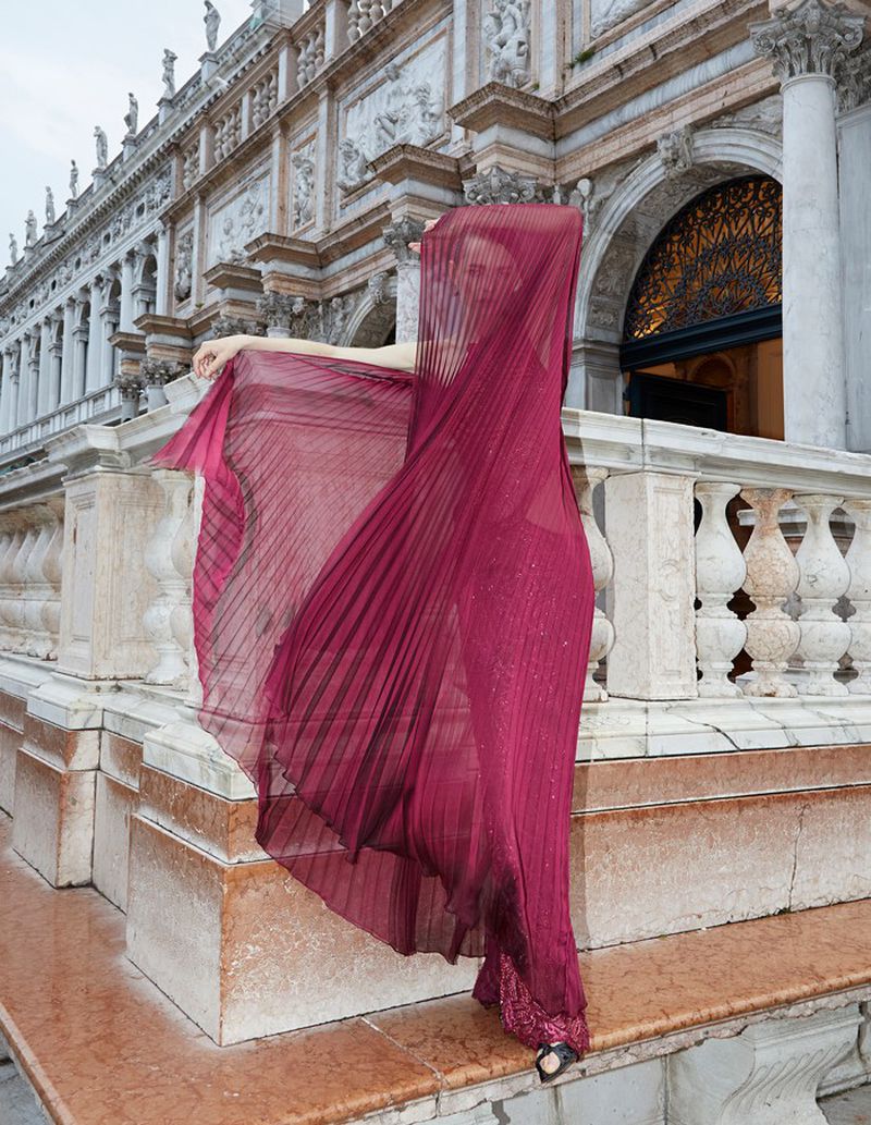 A woman in a pleated maroon gown stands on marble steps, partially veiled by fabric, with ornate architecture in the background.