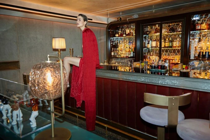 A woman in a red dress sits on a barstool in a dimly lit bar with a bar counter lined with bottles in the background.