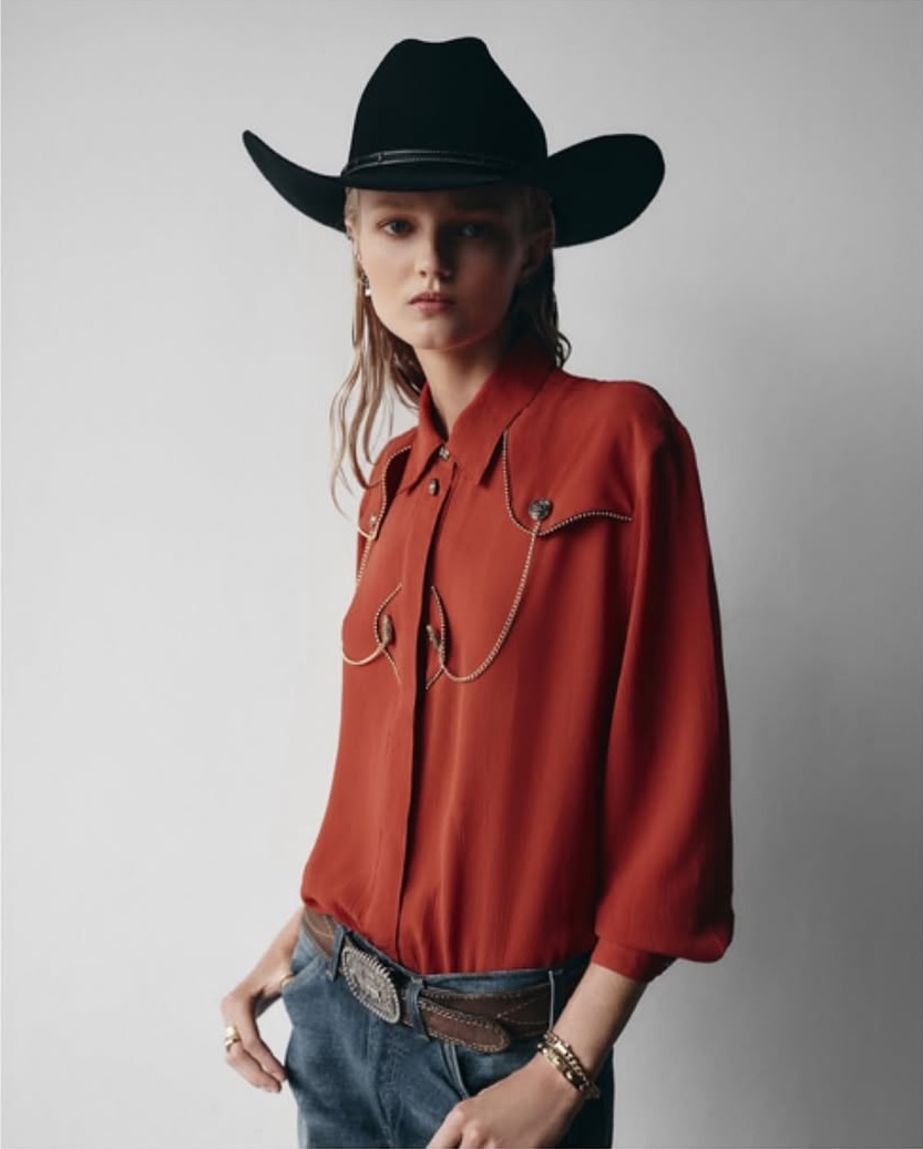 Blonde woman in a Western-style red shirt, jeans, and a black cowboy hat poses against a white background in a studio shot.