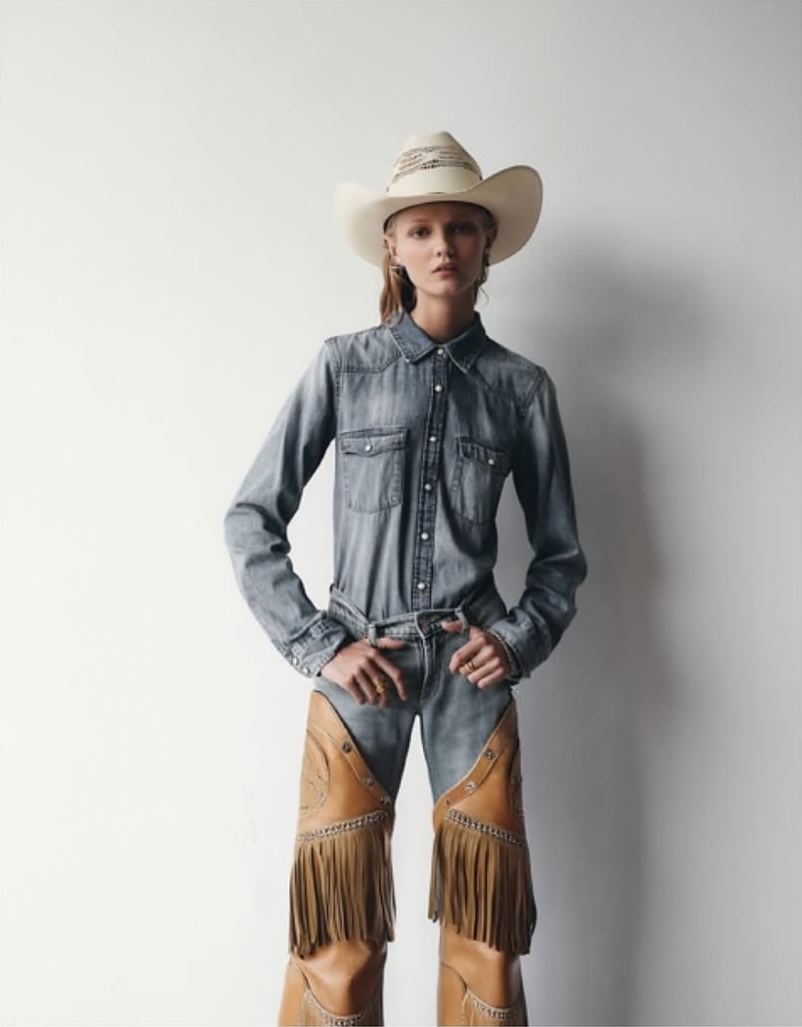 Woman in denim jumpsuit and chaps posing against a white wall, wearing a cowboy hat, fashion editorial shot, studio lighting.
