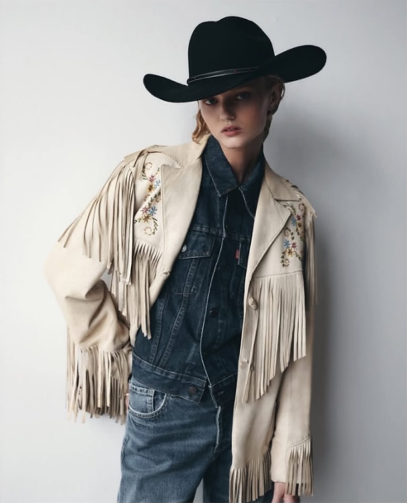A woman poses indoors wearing a black cowboy hat, denim jeans and jacket, and a light beige fringed jacket with embroidered details.