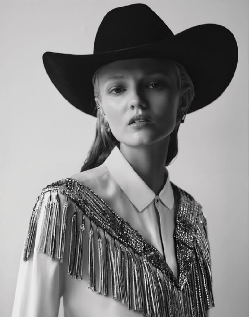 Monochrome portrait of a young woman wearing a cowboy hat and decorative fringe blouse looking at the camera in a photo studio.
