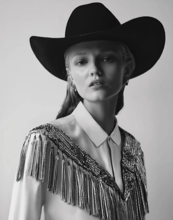 Monochrome portrait of a young woman wearing a cowboy hat and decorative fringe blouse looking at the camera in a photo studio.