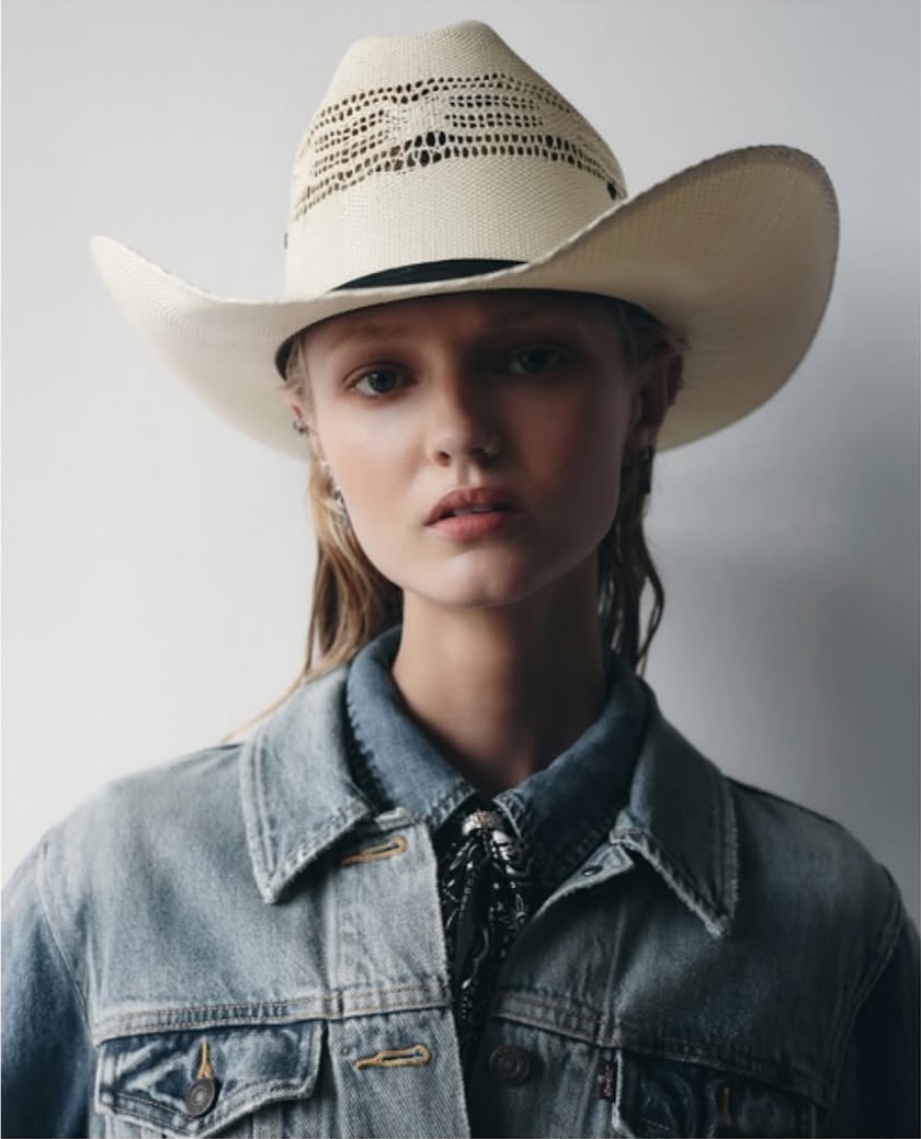 Portrait of a fair-skinned woman with a cowboy hat and denim jacket, looking directly at the camera against a neutral backdrop.
