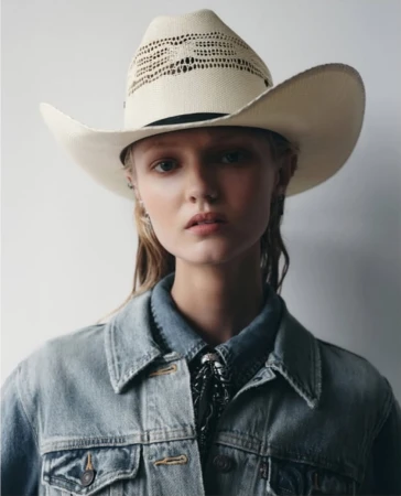 Portrait of a fair-skinned woman with a cowboy hat and denim jacket, looking directly at the camera against a neutral backdrop.