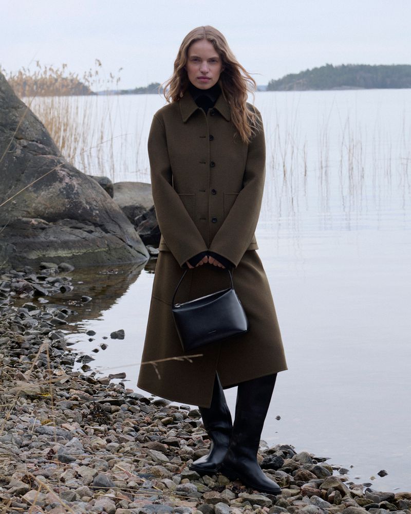 Adult woman poses in a green coat and skirt near a rocky lake shore with a black handbag and boots in an outdoor fashion shot.