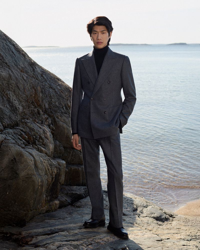 A stylish Asian man in a double-breasted suit poses near the ocean and a rocky formation on a bright day, creating a sophisticated outdoor portrait.