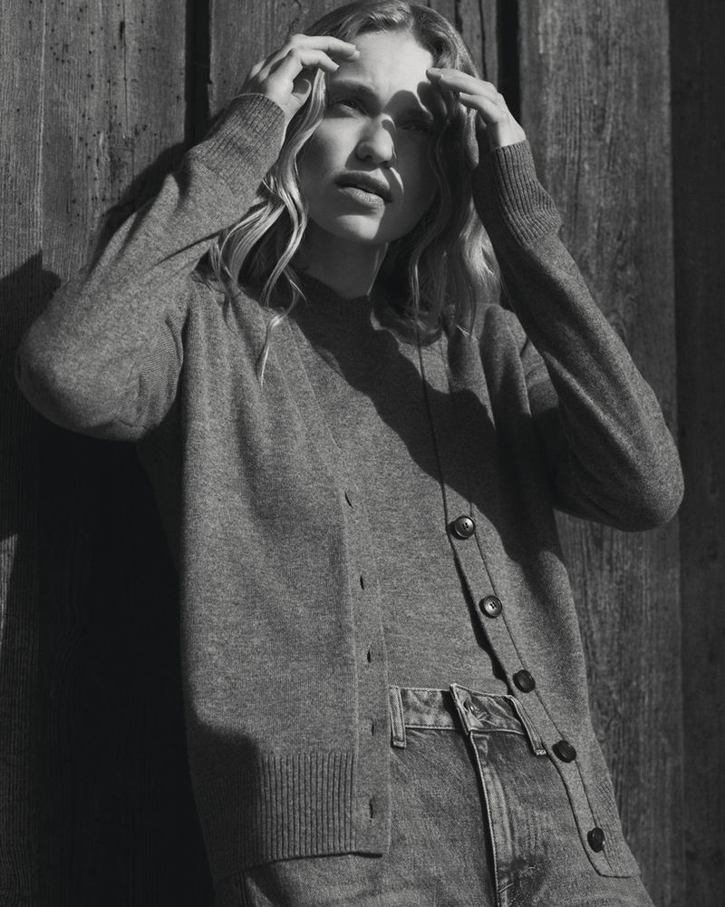 A young woman with blonde hair poses against a wooden wall, wearing a grey cardigan and denim jeans in a monochrome photograph.