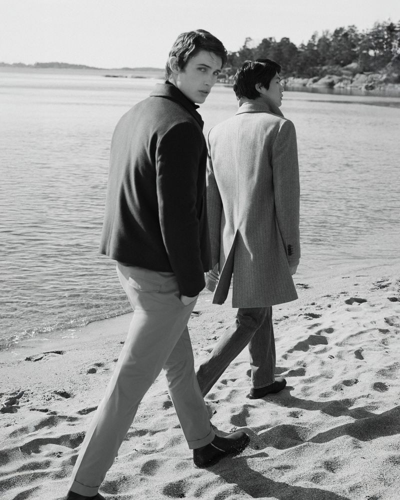 Two men walk along a sandy beach near the water's edge. One man looks back toward the camera, wearing a jacket and pants.