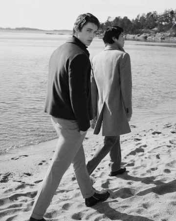 Two men walk along a sandy beach near the water's edge. One man looks back toward the camera, wearing a jacket and pants.