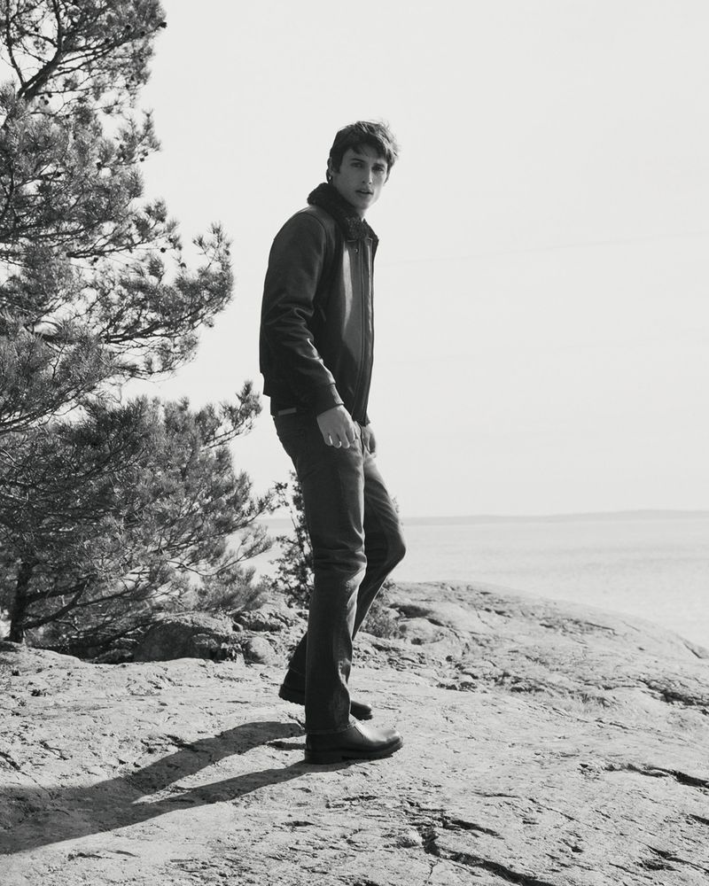 Monochrome portrait of a male model in a leather jacket and jeans standing on a rocky outcrop with trees and water in background.