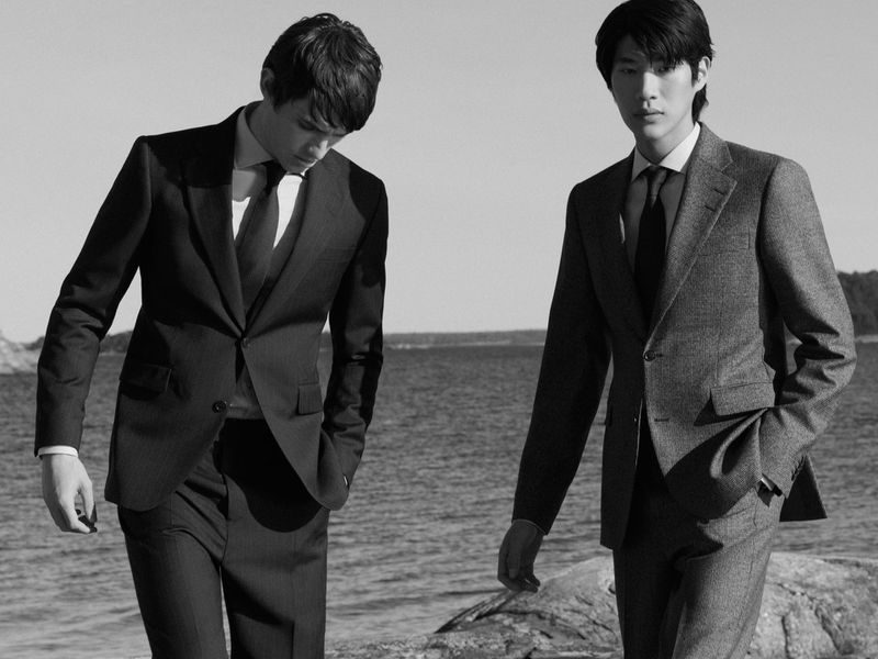 Black and white shot of two adult men in suits standing near a body of water, rocks, and trees on a clear day in a medium shot.