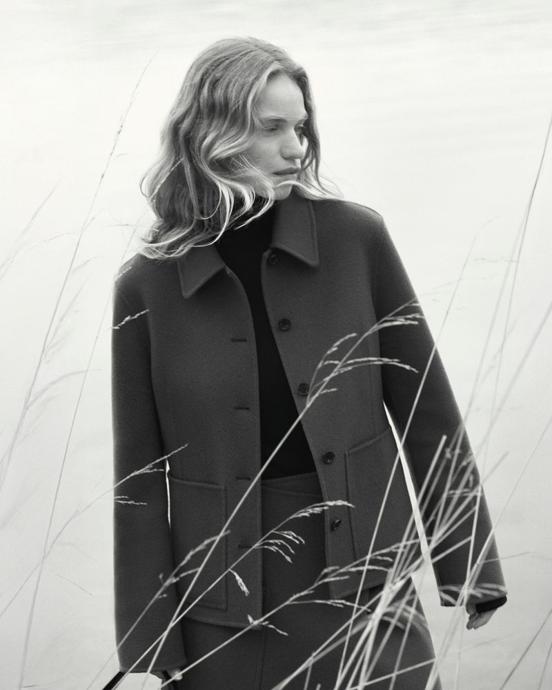 Monochrome medium shot of a woman in a jacket, gazing off-camera, standing among tall grasses in an outdoor setting.