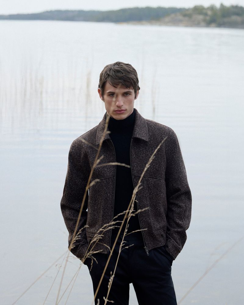 Adult male model posing outdoors near lake wearing brown jacket and black turtleneck in a fashion editorial shot, portrait orientation.