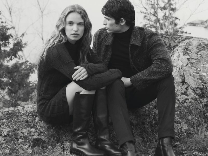 A young adult couple poses outdoors in black sweaters and boots. The woman has wavy blonde hair, and the man looks away from the camera.