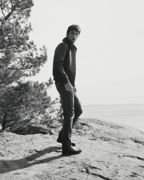 Monochrome portrait of a male model standing on rocky coast by the ocean wearing a dark jacket