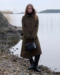 Model standing on a rocky lakeshore wearing an olive green coat and holding a black handbag.