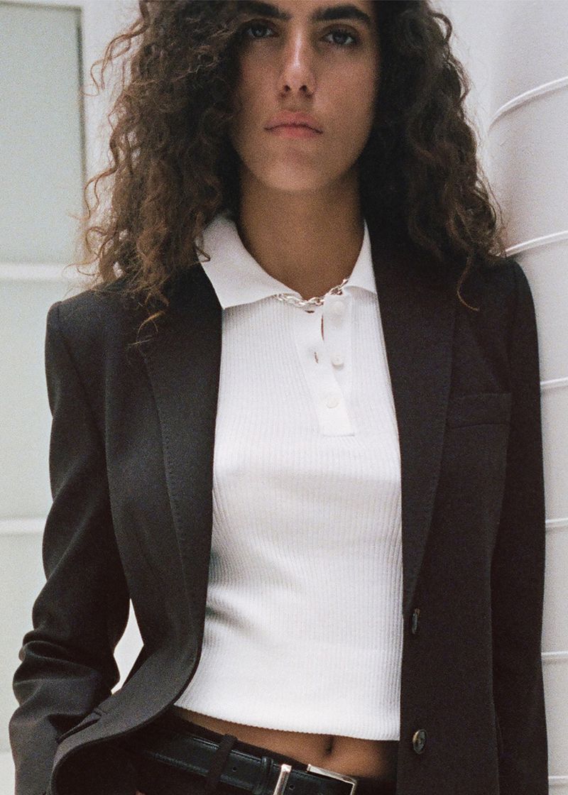 Brunette woman in a polo shirt and blazer poses indoors with neutral expression, highlighted by bright, even lighting and a simple background.