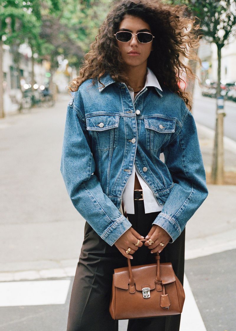 A stylish woman wearing a denim jacket and sunglasses crosses a city street, holding a brown leather satchel, with trees in the background.