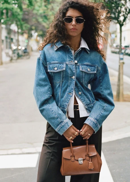 A stylish woman wearing a denim jacket and sunglasses crosses a city street, holding a brown leather satchel, with trees in the background.