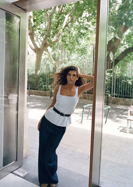 A young adult woman with curly brown hair poses in an doorway wearing a white tank top and wide-leg dark denim pants, outdoors.