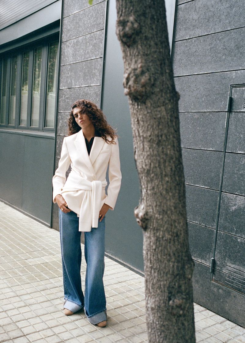 Fashion shot of an adult woman with curly hair posing in a white blazer and jeans next to a tree on a city street.