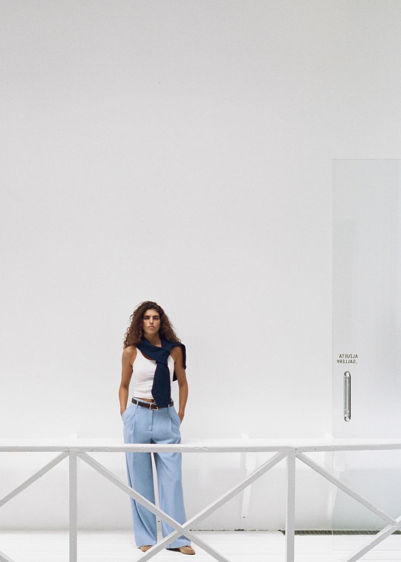Woman in a white tank top and light blue pants, standing against a white wall, with a navy blue sweater draped over her shoulders.