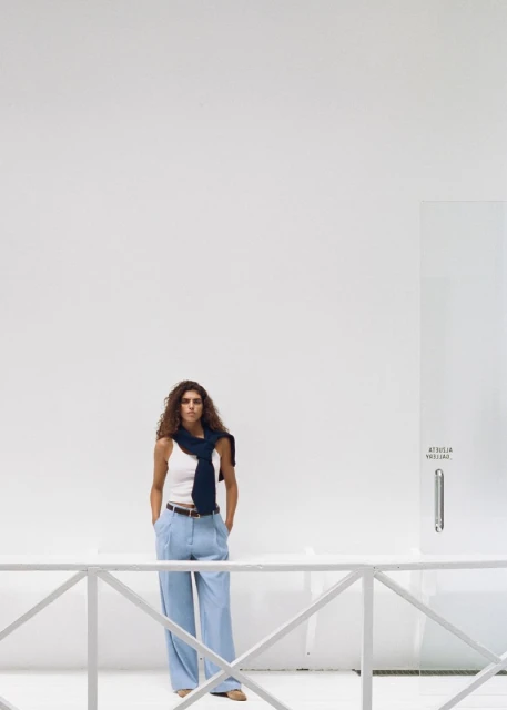 Woman in a white tank top and light blue pants, standing against a white wall, with a navy blue sweater draped over her shoulders.